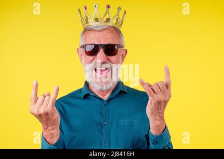 uomo anziano che guarda la macchina fotografica, che indossa una camicia di cotone verde con un colletto, corona d'oro e occhiali da sole in studio giallo background.mostrando il movimento dei rockers Foto Stock