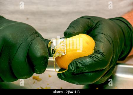 Il processo di preparazione di una bevanda alcolica Limoncello a casa. Mani maschili in guanti neri sbucciare la scorza da un limone. Foto Stock