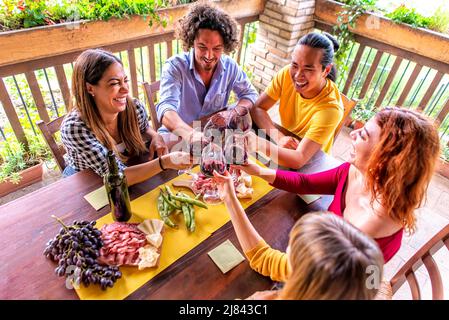 felice gruppo di amici multiratial fare un brindisi celebrativo con vino su un tavolo da picnic all'aperto in terrazza. persone che si riuniscono in un estate felice Foto Stock