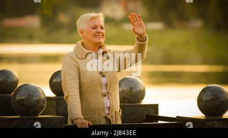 Ritratto di sorridente anziana bella femmina guardando via ondeggiante ciao o Arrivederci rilassarsi all'aperto in clima fresco, felice anziana donna grigio-capelli Foto Stock