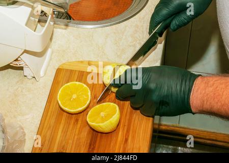 Il processo di preparazione di una bevanda alcolica Limoncello a casa. Le mani maschili in guanti neri tagliano un limone su un asse di legno con un coltello. Foto Stock