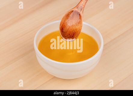 L'olio di girasole scorre da un cucchiaio di legno in una ciotola bianca su uno sfondo di legno. Spazio di copia. Foto Stock