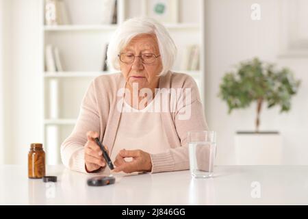 Donna anziana che controlla il livello di zucchero nel sangue e seduto a un tavolo con una bottiglia di pillole Foto Stock