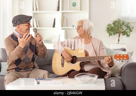 Uomo anziano felice con un microfono a casa e una donna anziana che suona una chitarra acustica seduta su un divano Foto Stock