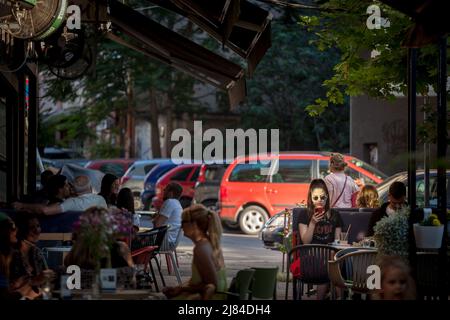 Foto di una terrazza piena nel centro della città di Belgrado, Serbia, con una donna che controlla il suo telefono seduto in un caffè, mentre altre persone sono celebrate Foto Stock