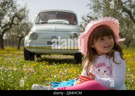 Foto di una sorridente bambina in un cappello seduto in un prato fiorito con un'auto d'epoca sullo sfondo durante un viaggio in Italia Foto Stock