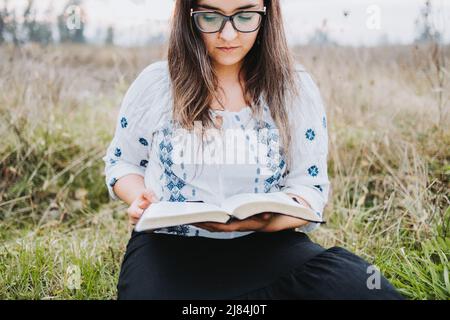 Donna con bicchieri seduti all'aperto sull'erba tenendo e leggendo una Bibbia aperta. Messa a fuoco selettiva. Foto Stock