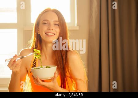 ragazza zenzero rossa in arancione sportivo top reggiseno mangiare salat in serata luce solare sfondo Foto Stock