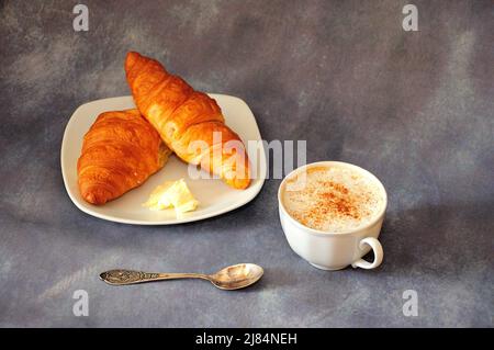 Una tazza di cappuccino alla cannella fresco e un piatto con due croissant e burro. Primo piano. Foto Stock