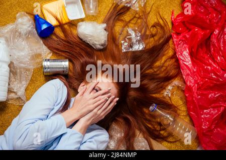 malcontento ragazza zenzero redhaired che ordina i rifiuti: bottiglia, carta e plastica che si stendono giù al tappeto del pavimento e piangendo Foto Stock