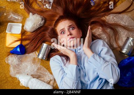 malcontento ragazza zenzero redhaired che ordina i rifiuti: bottiglia, carta e plastica che si stendono giù al tappeto del pavimento e piangendo Foto Stock