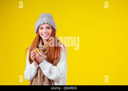 malattia malata donna zenzero rosso bello vestendo maglione a maglia e cappello con sciarpa bere tazza di tè caldo con limone in studio giallo Foto Stock