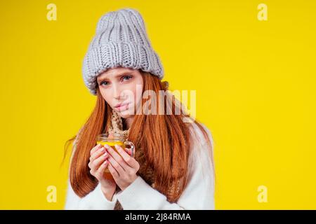 malattia malata rosso zenzero donna cattivo aspetto indossare maglione a maglia e cappello con sciarpa bere tazza di tè caldo con limone in studio sfondo giallo Foto Stock