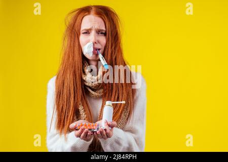 malbene rosso zenzero donna naso runny indossare maglione a maglia e sciarpa con molte pillole in mano in studio sfondo giallo Foto Stock