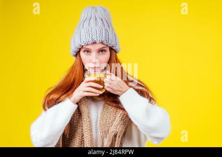 malattia malata rosso zenzero donna cattivo aspetto indossare maglione a maglia e cappello con sciarpa bere tazza di tè caldo con limone in studio sfondo giallo Foto Stock