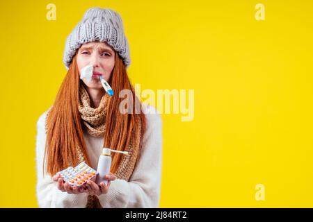 malbene rosso zenzero donna naso runny indossare maglione a maglia e sciarpa con molte pillole in mano in studio sfondo giallo Foto Stock