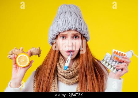 malattia malata donna rossa indossando maglione a maglia, cappello con sciarpa scelta tra limone naturale e zenzero rimedio popolare per raffreddori e mucchio di pillole Foto Stock