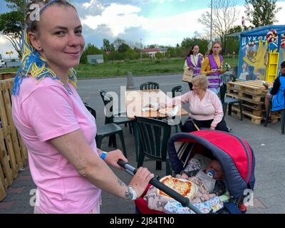 Przemysl, Polonia. 12th maggio 2022. Una giovane donna di Kharkiv dilaniata dalla guerra con trecce di mais decorate in sorrisi gialli e blu al suo bambino, o la pizza al suo giro sta per mangiare, dopo un viaggio sgarbante attraverso l'Ucraina dilaniata dalla guerra alla sicurezza in Polonia. Madre e bambino sono al Centro umanitario Tesco il 12 maggio, presso la tenda Italpizza, che ha fornito over100.000 pizze ai rifugiati ucraini, agli operatori umanitari e a chiunque venga in cerca di pizza, vicino al confine con Medyka. (Credit Image: © Amy Katz/ZUMA Press Wire) Foto Stock