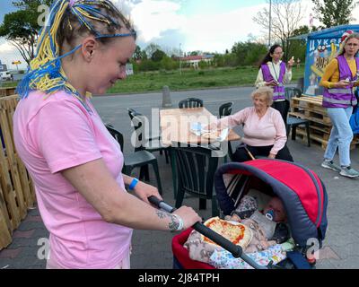 Przemysl, Polonia. 12th maggio 2022. Una giovane donna di Kharkiv dilaniata dalla guerra con trecce di mais decorate in sorrisi gialli e blu al suo bambino, o la pizza al suo giro sta per mangiare, dopo un viaggio sgarbante attraverso l'Ucraina dilaniata dalla guerra alla sicurezza in Polonia. Madre e bambino sono al Centro umanitario Tesco il 12 maggio, presso la tenda Italpizza, che ha fornito over100.000 pizze ai rifugiati ucraini, agli operatori umanitari e a chiunque venga in cerca di pizza, vicino al confine con Medyka. (Credit Image: © Amy Katz/ZUMA Press Wire) Foto Stock