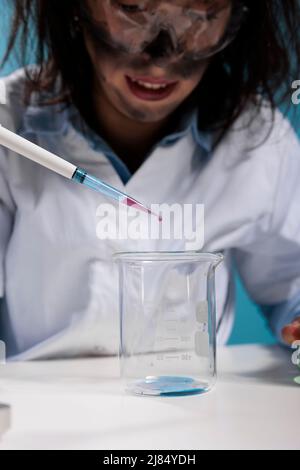 Primo piano di un biochimico maniacale dall'aspetto sciocco lunatico che utilizza il contagocce per sperimentare composti chimici tossici. Un operatore di laboratorio pazzesco che gocciola gocce di sangue in un bicchiere di vetro durante l'esecuzione di test pericolosi. Foto Stock