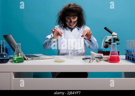 Pazzo esperto di biochimica stupida che mescola i composti chimici sperimentali usando le provette di vetro mentre si siede alla scrivania in laboratorio. Scienziato maniaco dall'aspetto ricercatissimo con sostanze che miscelano il viso spaventose. Foto Stock