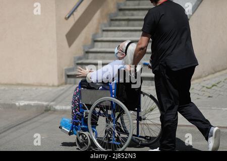 Bucarest, Romania - 9 maggio 2022: L'uomo ordinato spinge un paziente maschile anziano in una sedia a rotelle sul cemento nella corte di un ospedale rumeno. Foto Stock