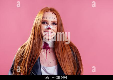 donna biker zenzero rossetto indossando giacca di pelle nera con sangue trucco chirurgia naso e collo frattura per halloween party, studio sfondo rosa Foto Stock