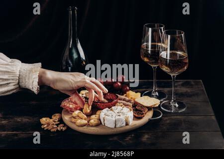 La mano della donna prende i cracker dal bordo degli antipasti con formaggio assortito, carne, rosette di salsiccia, uva e biscotti. Piatto di salumi e formaggi. Foto Stock