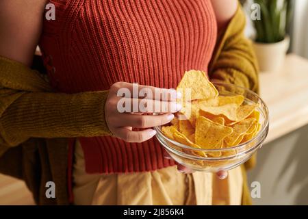 Primo piano di donna irriconoscibile in cima brillante e cardigan mangiare nachos da ciotola a casa, cibo malsano concetto Foto Stock
