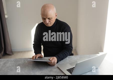 Confuso grigio capelli calvo 60 anni maschio in abiti formali di navigazione moderno tablet con bewilderment seduta in poltrona al tavolo Foto Stock