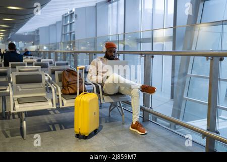 Giovane uomo nero concentrato seduto al terminal dell'aeroporto utilizzando un computer portatile indossare cuffie e ascoltare musica Foto Stock