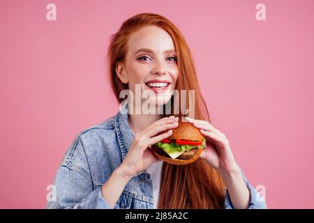 Donna zenzero rossa con greckles godendo di grande enorme cutlet hamburger e indossando giacca di jeans americano demino in studio sfondo rosa .USA Foto Stock