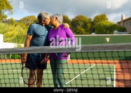 Coppia anziana amante del biraciale che tiene racchette baciare mentre si alza in piedi nel campo da tennis contro il cielo Foto Stock
