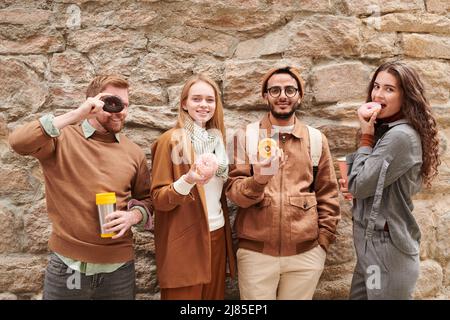 Gruppo di giovani e allegri turisti multietnici in abiti alla moda in piedi contro muro di pietra e mangiare ciambelle Foto Stock