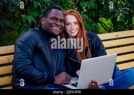 International Education Concept.Dark uomo con pelle wuth donna zenzero rossa guardando il laptop e seduto su panca, verde foglie sfondo Foto Stock