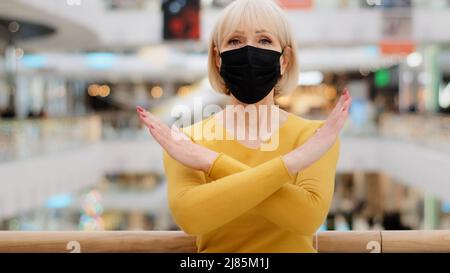 Donna matura gravemente sconvolto in maschera medica si erge in luogo pubblico guardando le braccia della telecamera incrociate di fronte rende proibire il gesto impone restrizioni Foto Stock