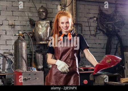 Forte e degna bella donna ginher rossa indossare casco di protezione e grembiule in pelle con guanti che reggono strumento saldatore . piccolo Foto Stock