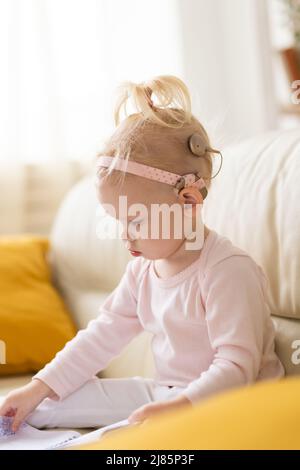 Bambino divertente con impianto cocleare seduto a casa. Ascolta il concetto di tecnologia innovativa di aiuto e medicina Foto Stock