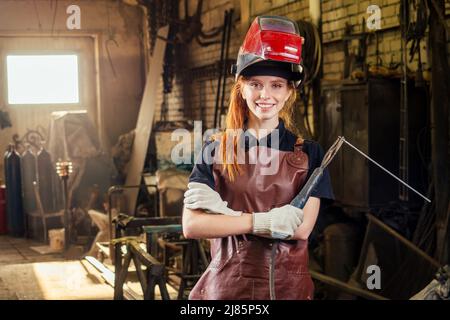 Forte e degna bella donna ginher rossa indossare casco di protezione e grembiule in pelle con guanti che reggono strumento saldatore . piccolo Foto Stock