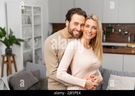 Felice coppia sposata in posa per una foto in cucina. I cuori più felici fanno le case più felici, il marito caucasico abbraccia la moglie da dietro. Concetto di relazioni armoniche Foto Stock