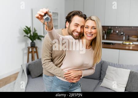 Felice coppia di famiglia caucasica che mostra nuove chiavi della casa alla macchina fotografica mentre posa in casa. Casa propria, proprietà immobiliare e alloggi. Ipoteca e acquisto di appartamento Foto Stock