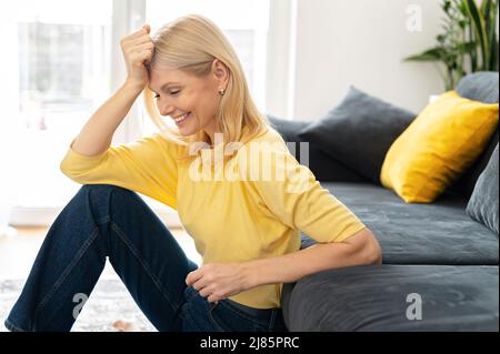 Donna anziana in casa interiore. Affascinante signora caucasica matura guardando giù e ridendo, riposandosi vicino al divano. Una donna serena trascorre il fine settimana a casa Foto Stock