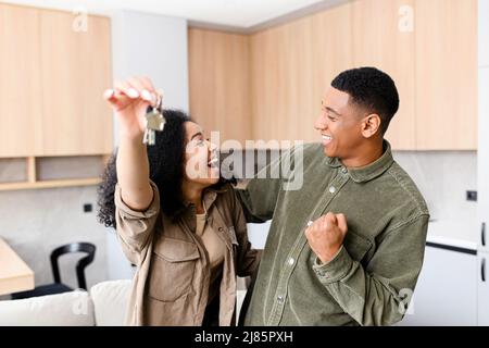 Donna multirazziale che tiene le chiavi di casa mentre abbraccia il ragazzo in un nuovo appartamento. Bella coppia sensazione estatica circa il loro recente acquisto di nuova casa Foto Stock