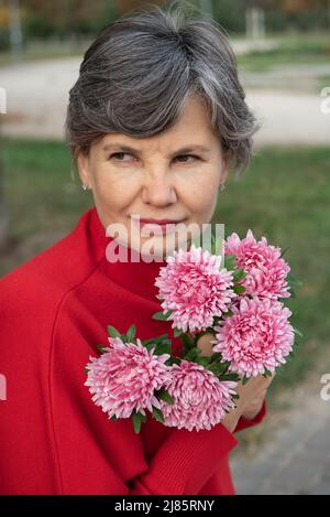 Ritratto semi-profilo di attraente donna dai capelli grigi in una blusa rossa con un bouquet di crisantemi viola, guardando da un lato. Girata in vita. Foto Stock