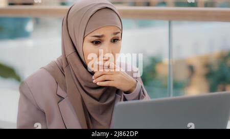 Triste musulmana ragazza studentessa in hijab guardando lo schermo del laptop che legge il email scioccato dalle notizie difettose frustrate le preoccupazioni della donna d'affari circa i problemi finanziari Foto Stock