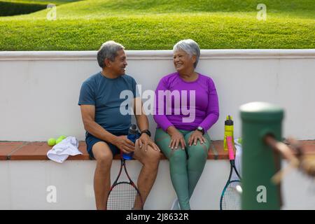 Coppia anziana felice biraciale che parla mentre si siede con racchette e bottiglie d'acqua su panca a corte Foto Stock