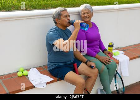 Sorridente donna anziana biraciale che guarda il marito che beve acqua mentre si siede sulla panca alla corte Foto Stock