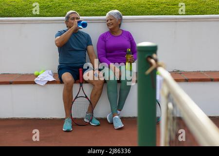 Tutta la lunghezza di stanco coppia anziana biraciale bere acqua mentre si siede su panchina al campo da tennis Foto Stock