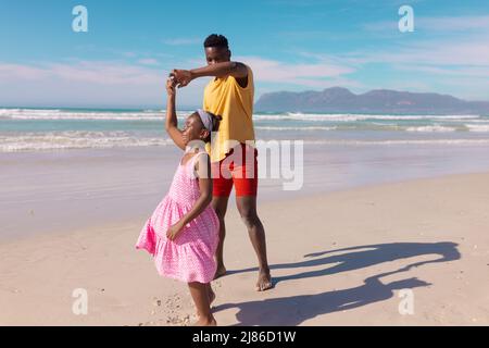 Giovane americano africano che balla con la figlia felice in spiaggia contro il mare e il cielo in giorno di sole Foto Stock