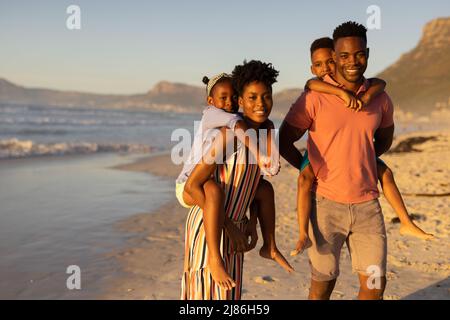 Ritratto di felice afroamericano giovani genitori piggybacking figlio e figlia in spiaggia contro il cielo Foto Stock